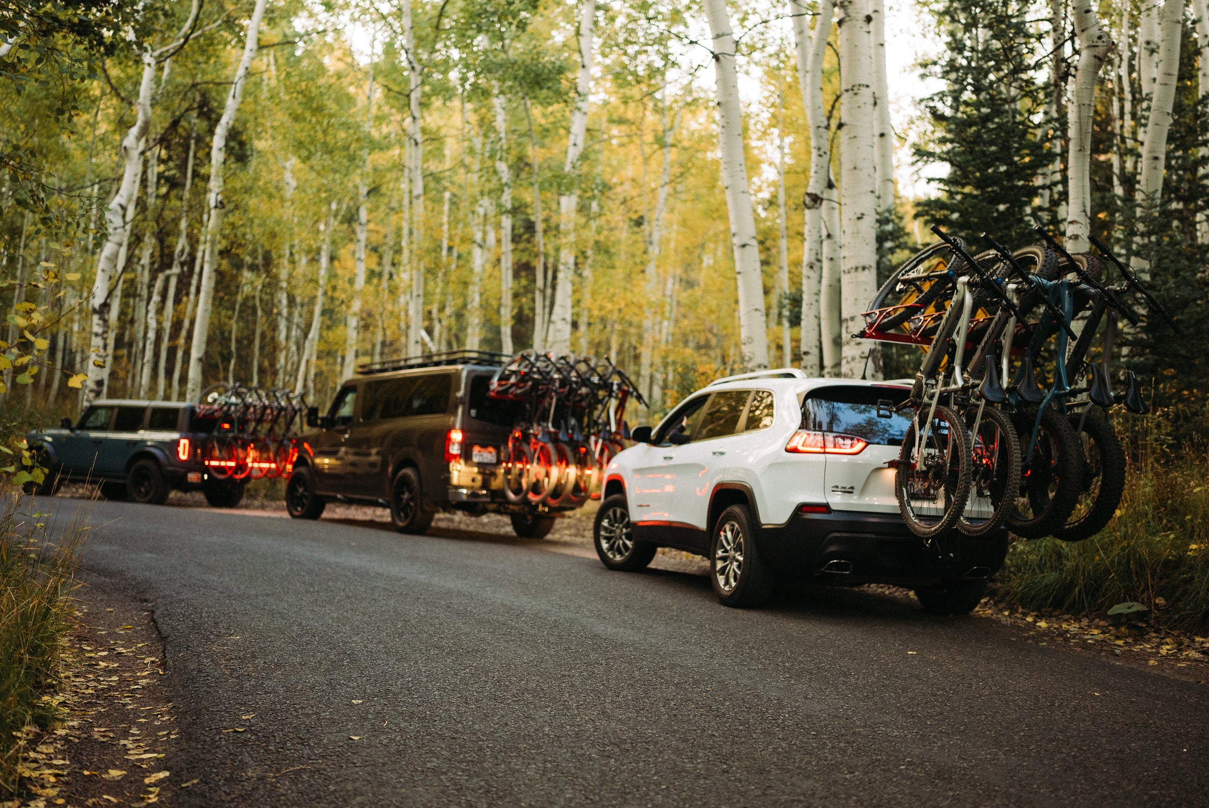 3 vehicles on a forest road with hitch mount vertical bikes racks on all of them, full of racked bikes.